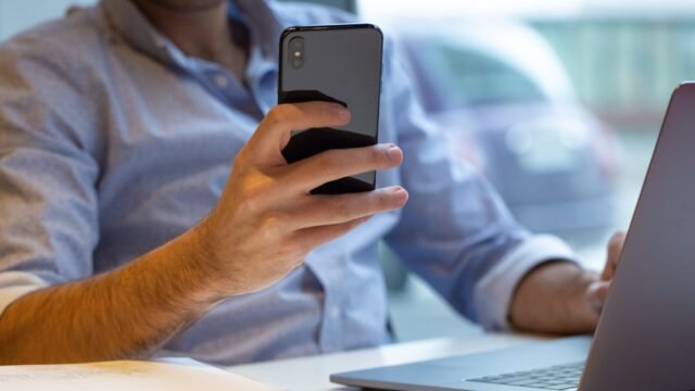 Homem de camisa social segurando o celular em frente a um notebook aberto