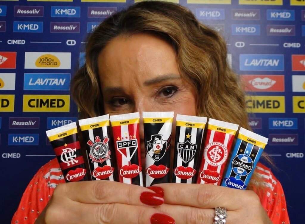 Mulher segurando tubos de hidratante labial Carmed, da Cimed, com escudos de clubes de futebol, em frente a painel com marcas da empresa