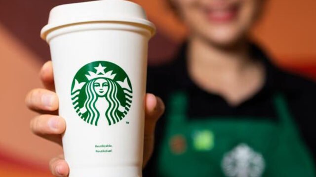 Mão segurando copo branco da Starbucks com o logotipo verde, com uma atendente de avental verde desfocada ao fundo